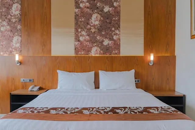 A made double bed with white pillows sits in front of a wooden headboard panel with floral wallpaper, flanked by wall lights and a bedside telephone.