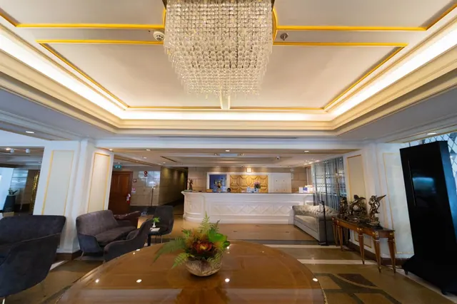 A hotel lobby with a crystal chandelier, a reception desk in the background, armchairs, a sofa, and a round table holding a floral arrangement.