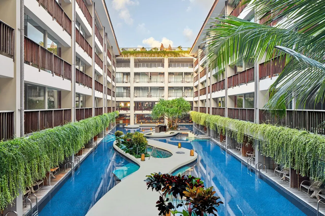 Two long blue pool channels run between multi-storey hotel buildings with balconies, separated by a curved white walkway and a planted island, with loungers along the edges.
