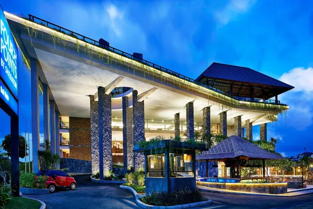 A hotel building exterior at dusk with an illuminated signboard, a covered driveway, stone pillars and a thatched pavilion.