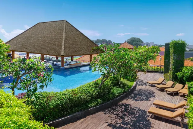 A large blue swimming pool sits beside a thatched roof pavilion, with sun loungers on a timber deck and frangipani trees around it.