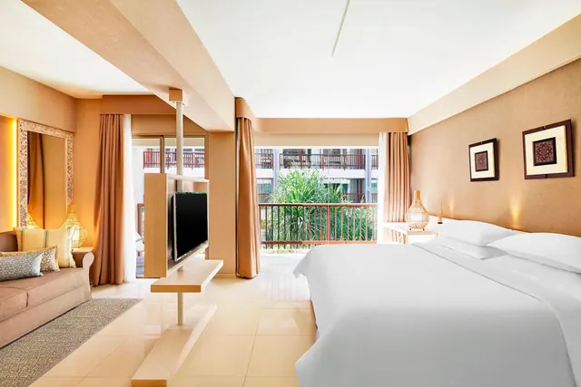 A bedroom with a large white bed, a sofa, a wall-mounted television and a glass door opening onto a balcony with railings and plants.