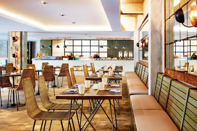 A dining room with wicker chairs and wooden tables set with white cups, a long banquette along the right wall, and a buffet counter in the background.