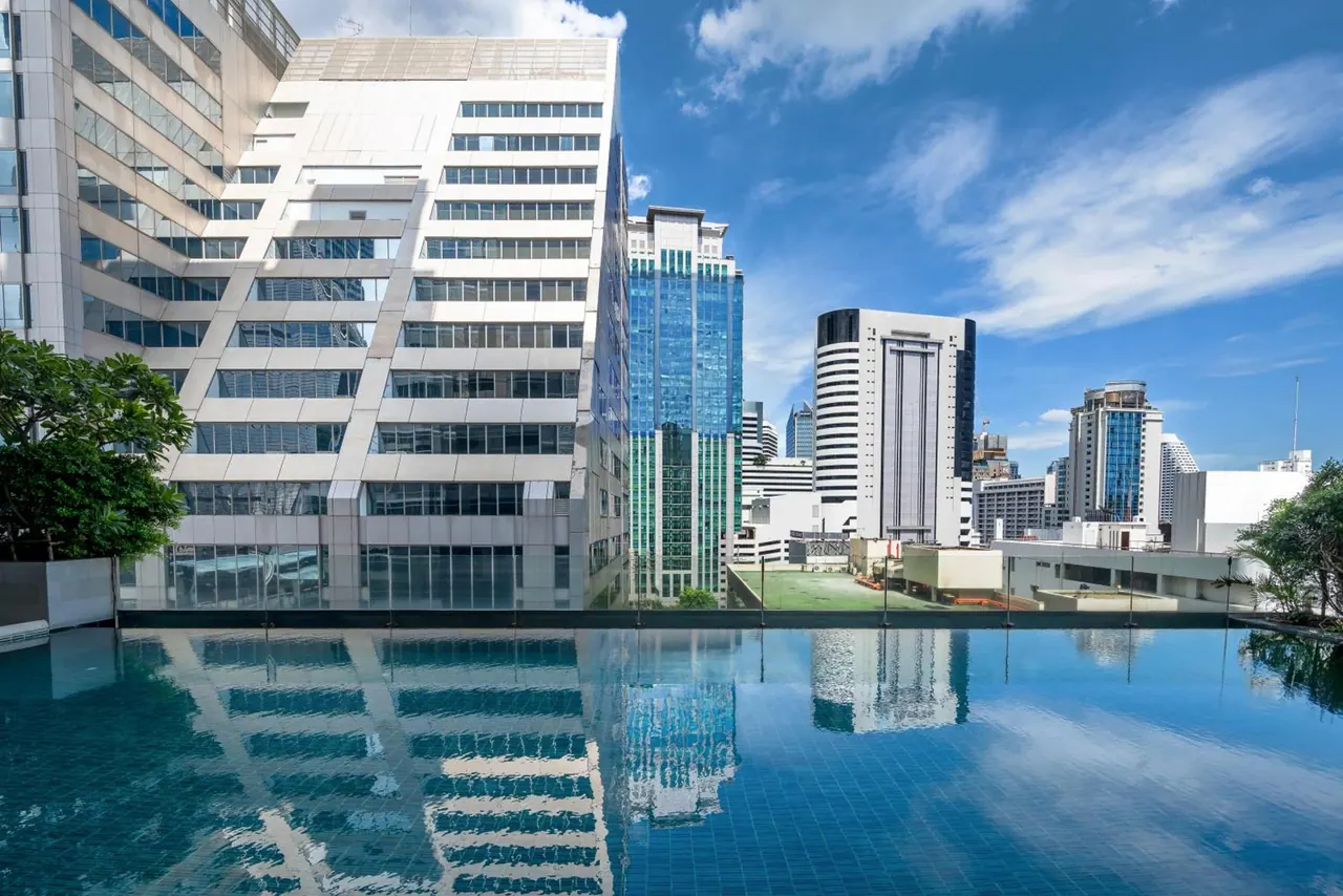 An infinity edge pool on a rooftop overlooks office and residential towers under a partly cloudy blue sky.