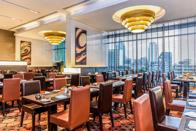 A restaurant dining room with rows of tables and leather chairs, tiered gold chandeliers, and tall windows looking out over city buildings.