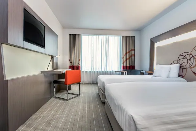 A hotel bedroom with two beds, a wall-mounted television, a desk with an orange chair, and a seating area near the window with sheer curtains.