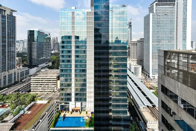 Elevated view of a glass high-rise with a pool and sun loungers at its base, surrounded by other city towers under a partly cloudy sky.