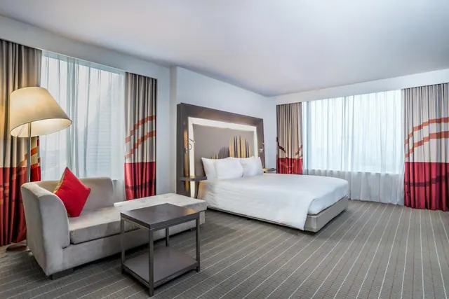 A bedroom with a king bed, a grey sofa with a red cushion, a floor lamp and a low side table, with large windows behind sheer curtains.