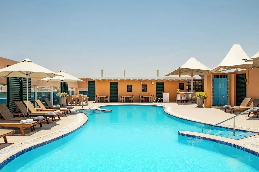 An outdoor swimming pool with sun loungers, white parasol umbrellas and tables with chairs beside an orange building under a clear sky.