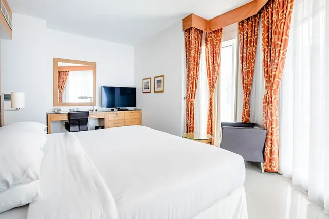 A bedroom with a large white bed, a desk with a mirror and television, an armchair, and orange patterned curtains beside tall windows with sheer drapes.