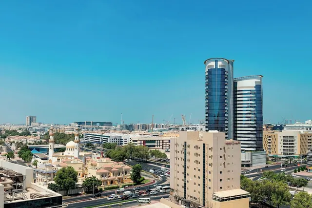 View from an elevated position over a city with tall glass towers, a domed mosque with minarets, low-rise buildings, a busy road and cranes under a clear blue sky.