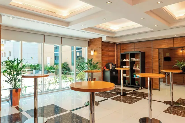 A lobby lounge with high round wooden-topped tables on a checkered marble floor, a coffee station with shelving, and floor-to-ceiling windows looking onto a courtyard with plants.