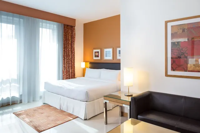 A hotel bedroom with a made double bed with white linen, a dark leather sofa, a glass side table with a lamp, and framed artwork on the walls.
