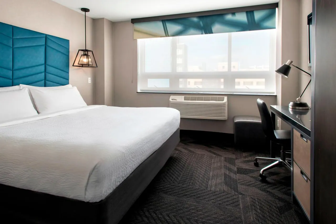 A bedroom with a king bed under a teal upholstered headboard, a desk with a lamp on the right, and a window looking out over city buildings with a wall air conditioner beneath it.