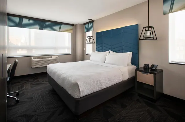 A bedroom with a king bed, a blue upholstered headboard, pendant lights, a work desk and a wall air conditioning unit under the window.
