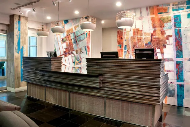 A reception desk clad in stacked wooden panels stands in a lobby beneath pendant lights, in front of a colourful abstract map-style mural.