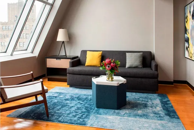A living area with a dark grey sofa, yellow and grey cushions, a white armchair, a blue hexagonal side table with flowers, and a view of a brick building through an angled window.
