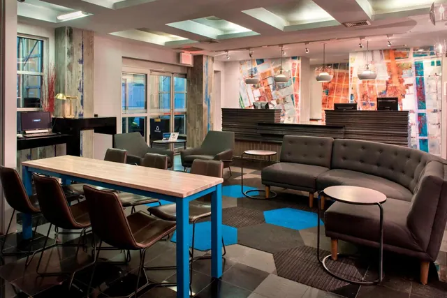 A lobby lounge with grey sofas, a high table with blue legs and brown chairs, and a reception desk in front of a multicolored wall mural.