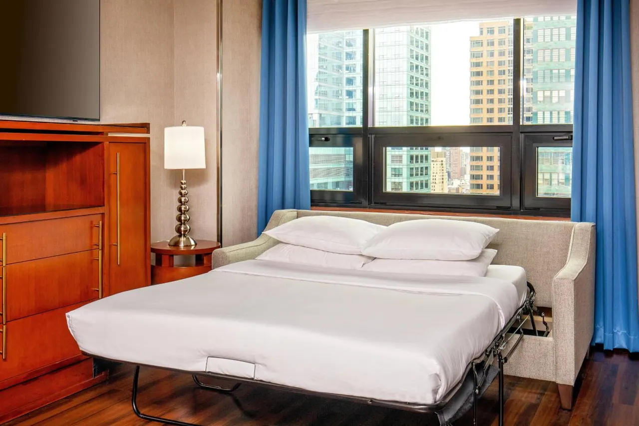 A bedroom with a made-up pull-out sofa bed, a wooden cabinet below a wall-mounted TV, blue curtains, and a window looking out over city buildings.