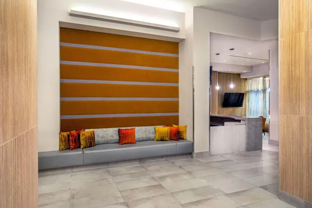 A lobby seating area with a long grey bench, an orange striped wall and colourful cushions, with a reception desk and wall-mounted television visible in the background.