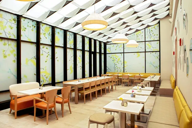 A bright dining room with a translucent fabric ceiling, pendant lamps, botanical-print glass walls, rows of tables and chairs, and yellow banquette seating.