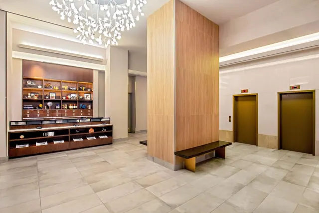 A lobby with a white branch-style chandelier, a wood-panelled column, a dark wooden bench, a display shelf and two elevator doors.