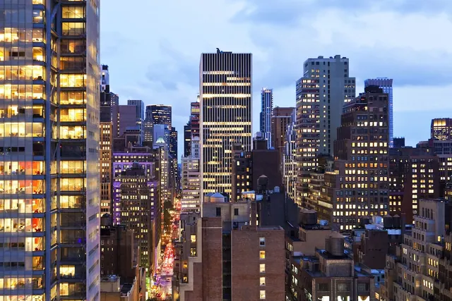 A dusk view over tall city buildings with many lit windows, with a busy street lined with traffic light trails visible below.