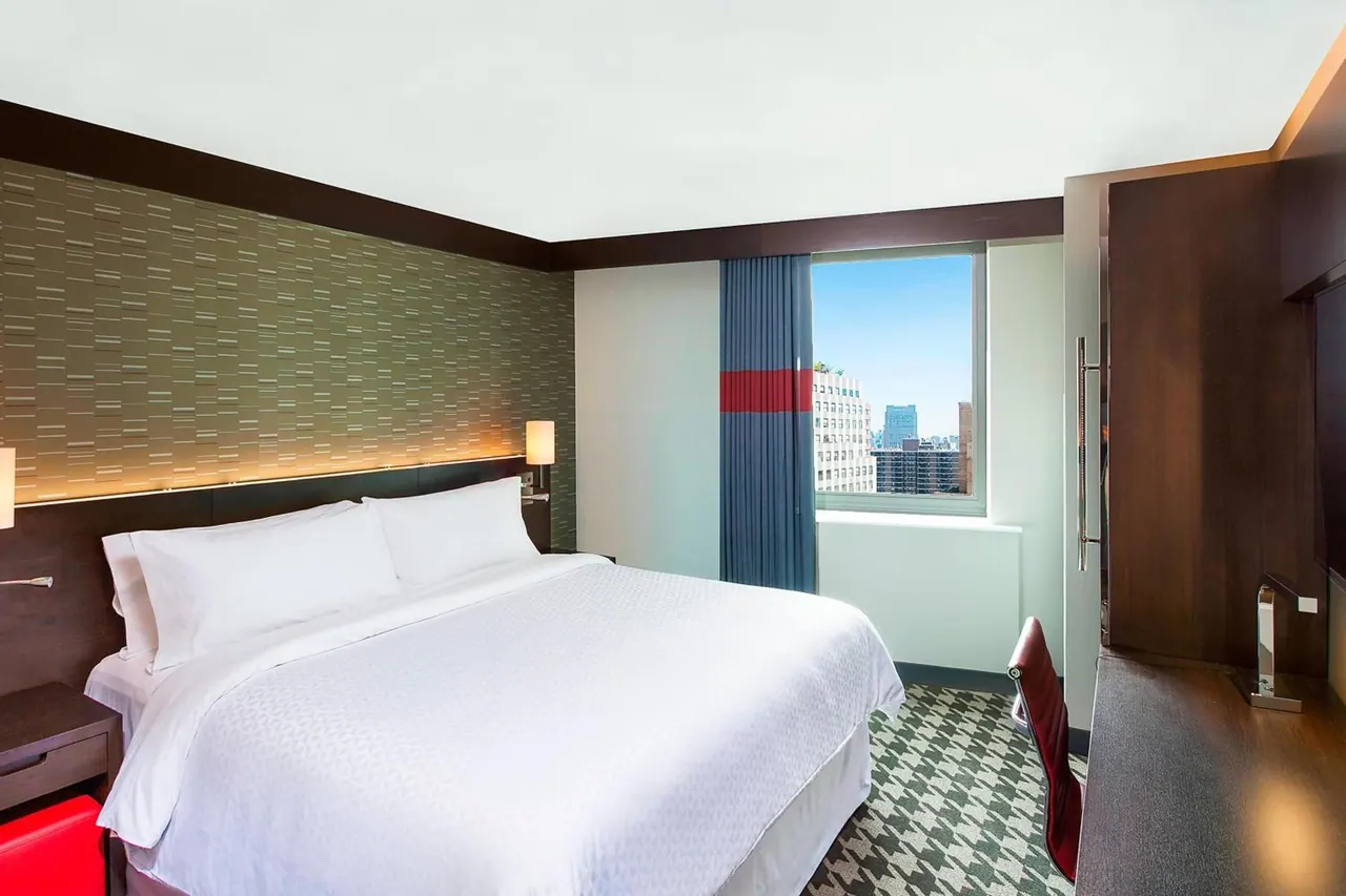 A king bed with white linens sits beside a window showing city buildings and blue sky, with a wood desk and wall-mounted television on the right.