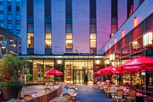 The exterior of a Four Points by Sheraton hotel at dusk, with an outdoor dining area of red umbrellas and bistro chairs in front.