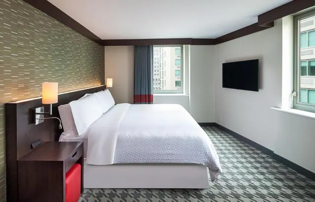 A bedroom with a made king bed, a wall-mounted television and a window looking onto nearby city buildings.