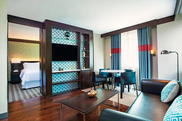 A living area in a hotel suite with a wall-mounted television in a dark wood unit, a coffee table, a round dining table with chairs, and a leather sofa, with a bedroom visible through the doorway on the left.