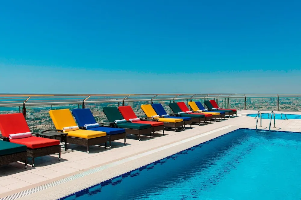 A rooftop pool with a row of colourful sun loungers, a small second pool at right, and a hazy city view beyond the glass railing.