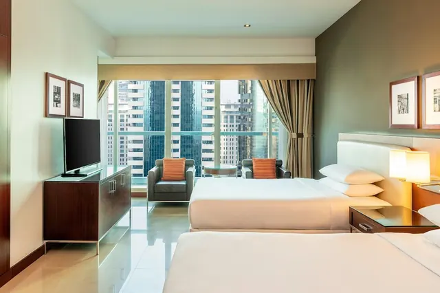 A hotel bedroom with two beds, a pair of armchairs by large windows overlooking high-rise towers, and a flat-screen television on a wooden sideboard.