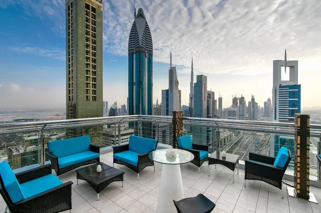 A terrace with blue cushioned wicker seating and glass tables overlooks a city skyline of high-rise towers, with the Burj Khalifa visible in the centre.