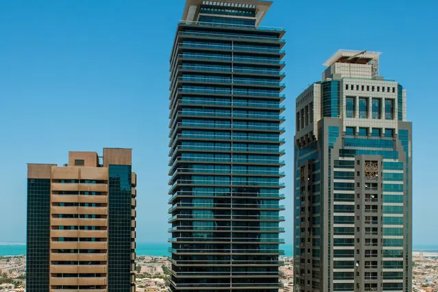 Three high-rise towers with glass facades stand against a clear blue sky, with the sea and low-rise city buildings visible below.