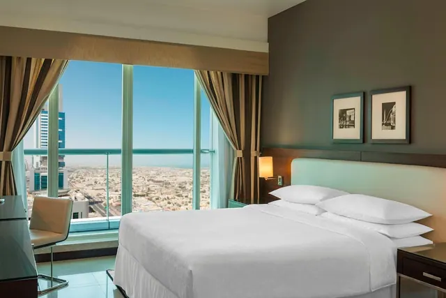 A bedroom with a made king bed, a desk and chair, and floor-to-ceiling windows looking out over a low-rise city under a clear sky.