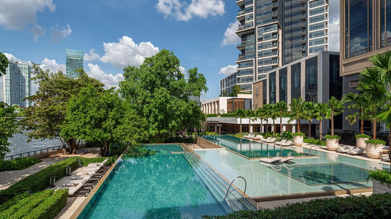 Outdoor swimming pool with sun loungers beside a tree-lined riverbank and high-rise towers under a partly cloudy sky.