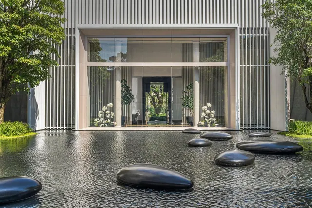 The hotel entrance has a glass facade with a glossy water feature in front of it, dark stones set in the water, and trees on either side.