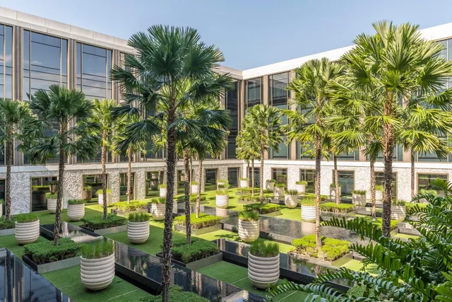A courtyard garden with rows of palm trees, large white planters and dark water channels, surrounded by a multi-storey building with glass facades.