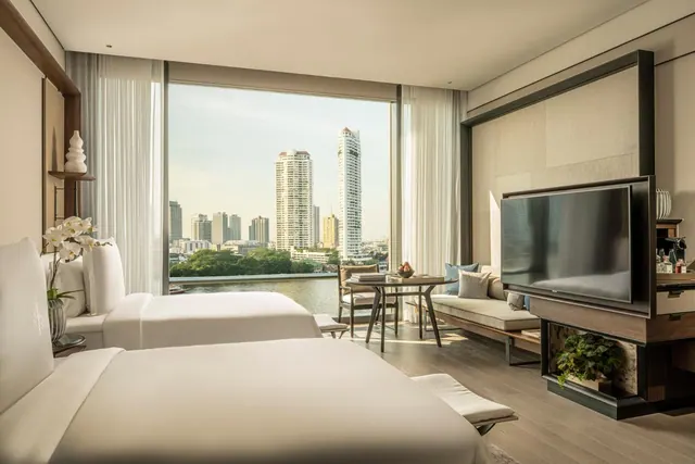 A hotel bedroom with two beds, a wall-mounted television, a seating area and floor-to-ceiling windows overlooking a river and city high-rises.