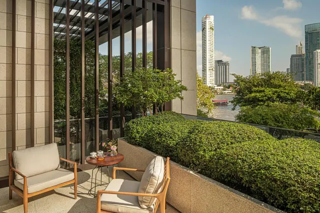 A private balcony with two armchairs and a small round table with tea items overlooks a river and a city skyline with tall towers.