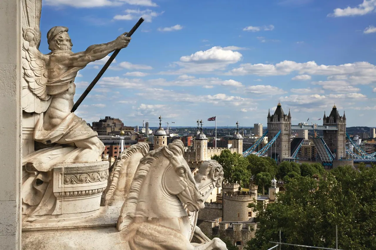 Stone sculptures of a winged figure with a spear and a horse in the foreground overlook Tower Bridge and the Tower of London under a blue sky with clouds.