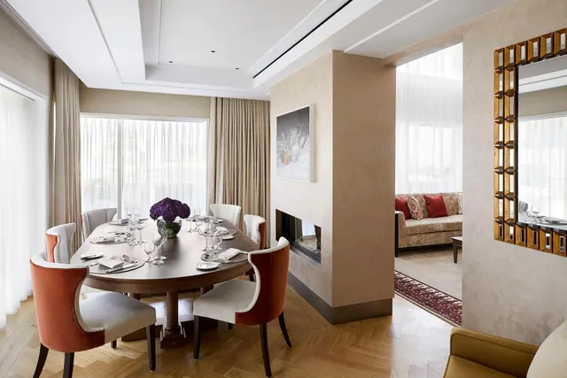 A dining table laid for a meal sits beside a fireplace, with a sofa and sheer-curtained windows visible in the adjoining living area.