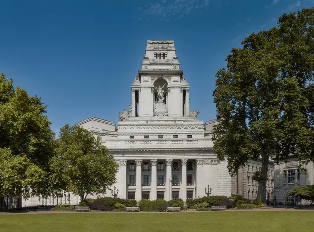 A pale stone classical building with a tall central tower and a columned front stands behind a lawn and benches under a clear blue sky.