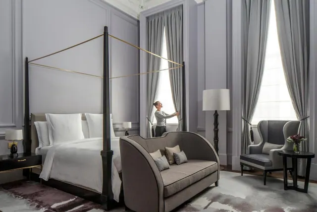 A bedroom with a four-poster bed, a sofa and two armchairs, with a staff member adjusting grey curtains at a tall window.