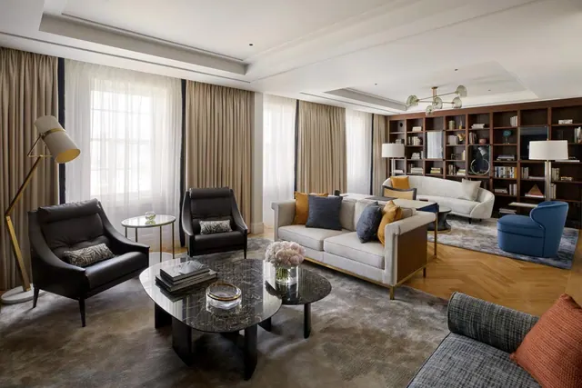A living room with two leather armchairs, a marble coffee table, a light grey sofa, and a wall of built-in bookshelves beside tall windows with sheer and lined curtains.