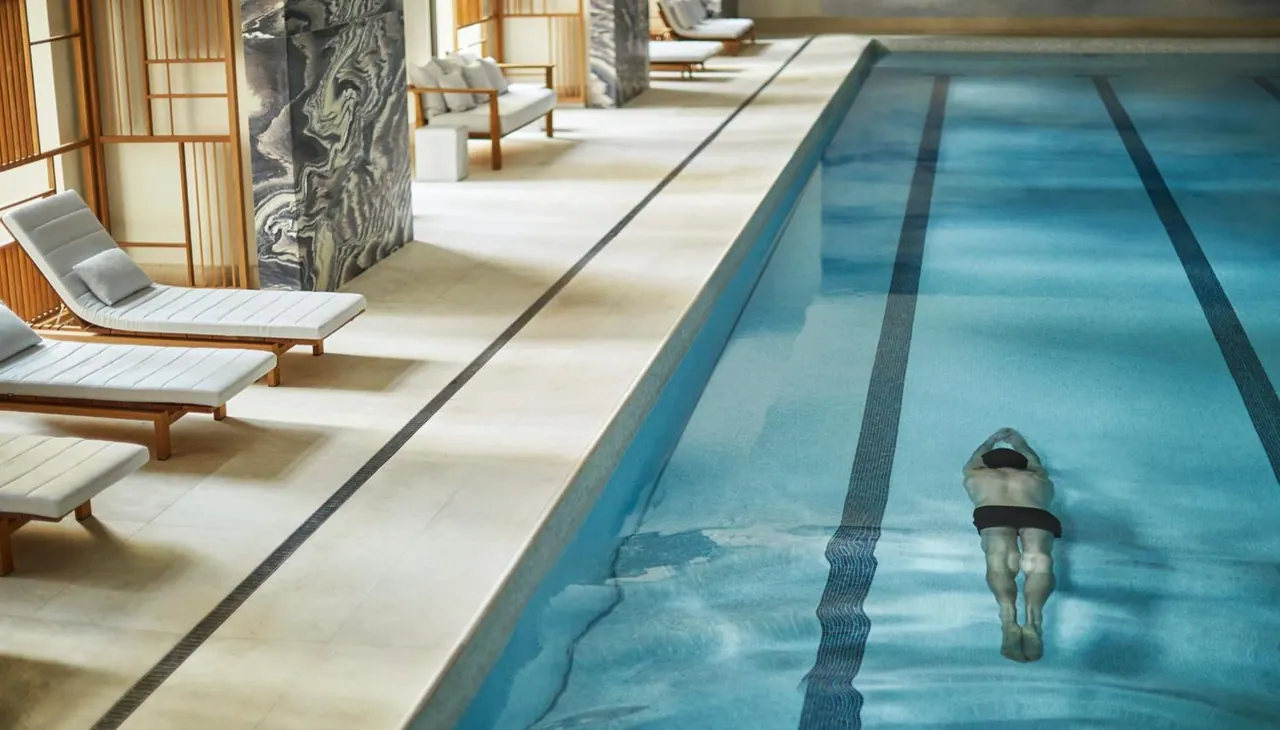 An indoor lap pool with lane lines and one swimmer, with white sun loungers along the side beneath wooden screens and dark marble columns.