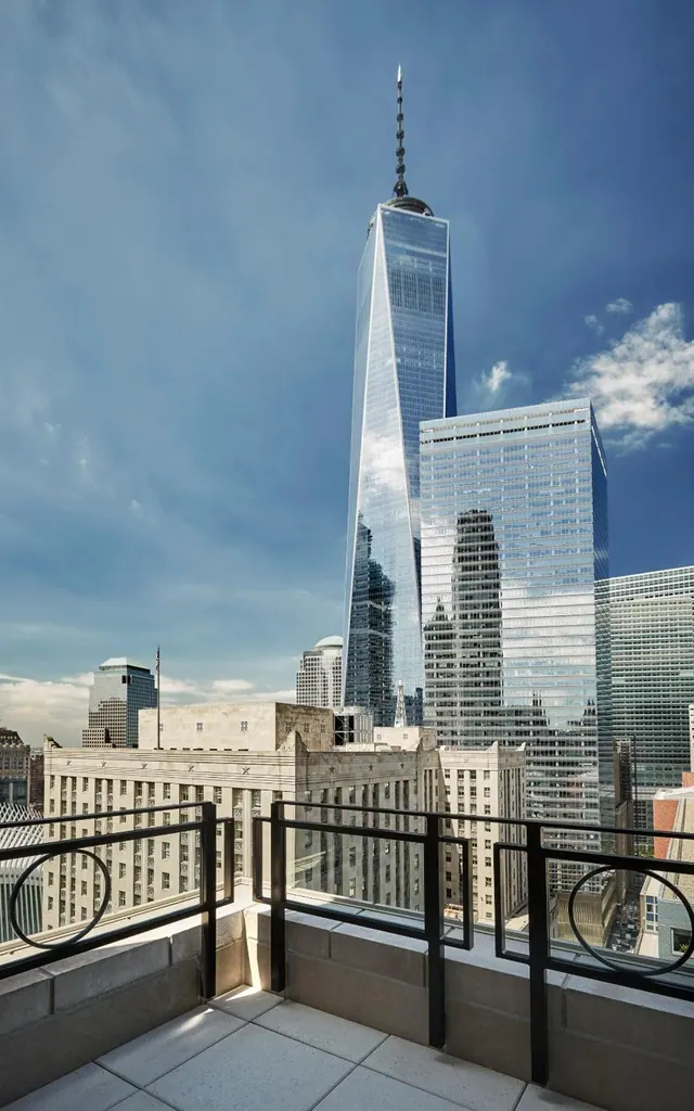 A stone terrace with black metal railings looks out over nearby buildings toward One World Trade Center under a blue sky.
