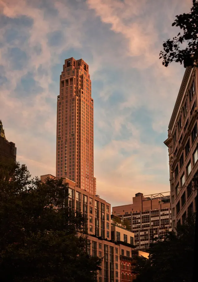 A tall terracotta-coloured skyscraper with stepped setbacks rises above lower buildings and trees in warm evening light.