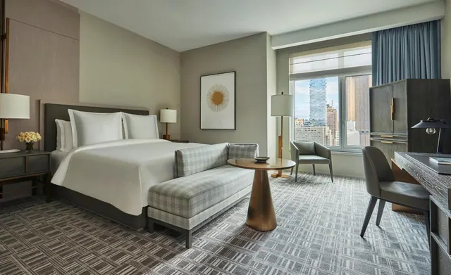 A king bed with a upholstered bench at its foot, a round side table, an armchair and a desk in a hotel room, with a window showing city skyscrapers.